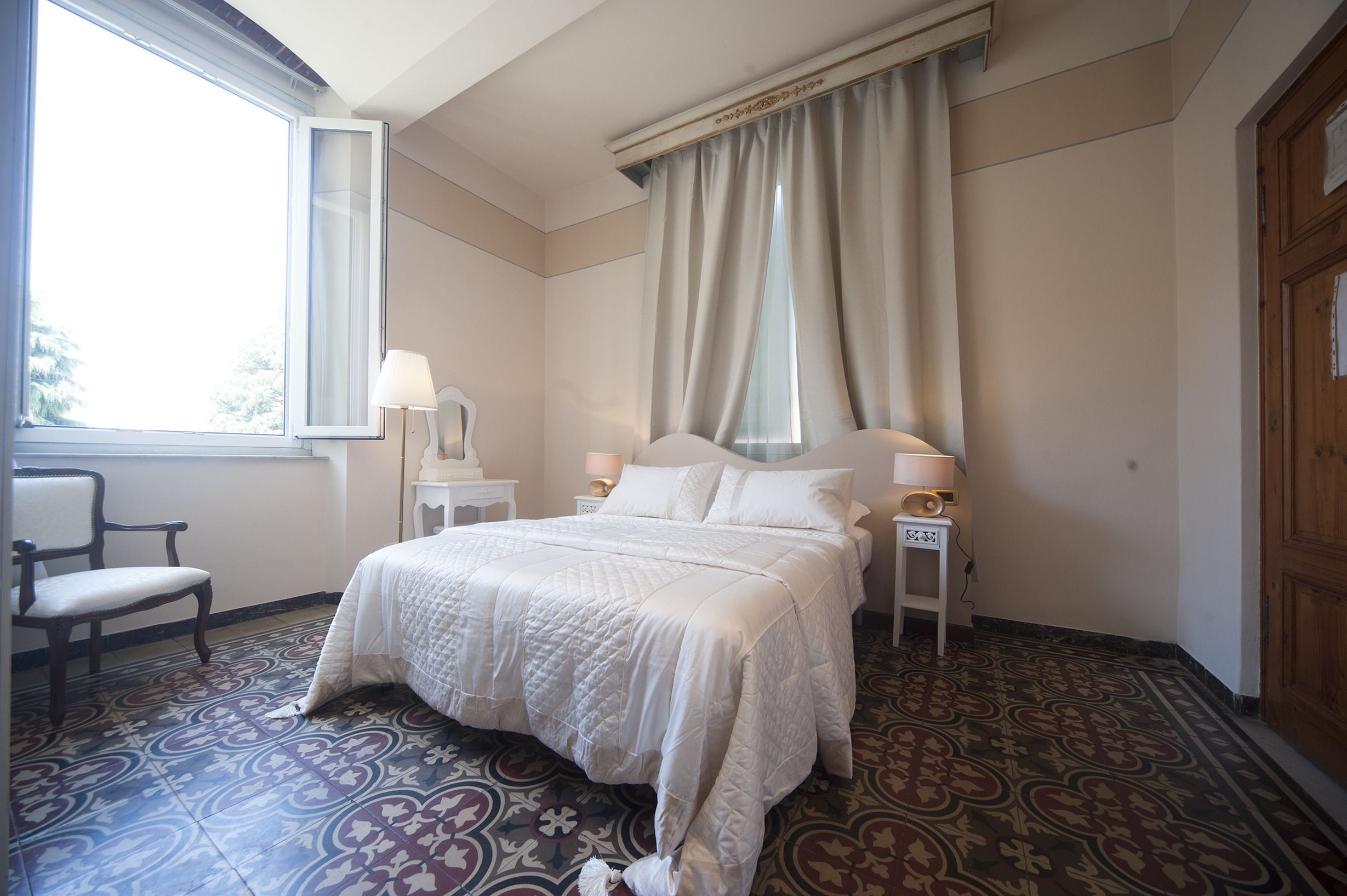 Quarto com cama grande, duas mesas de cabeceira com luminárias, uma cadeira, janela com cortinas e ladrilhos no chão com padrão.