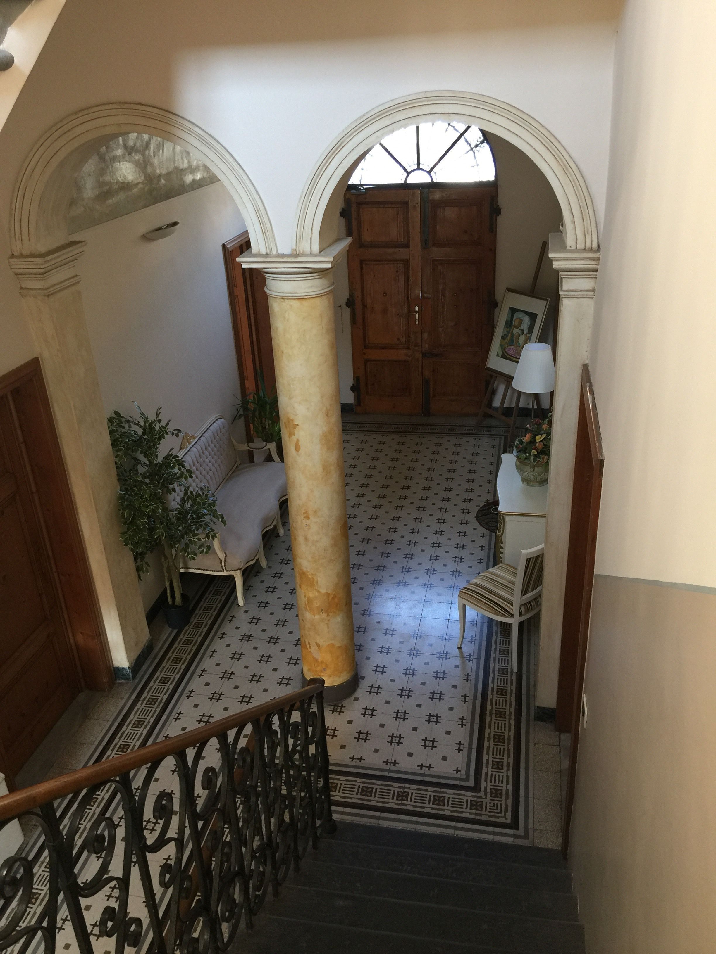Un escalier dont les marches sont décorées d’un motif particulier mène à une pièce dotée d’une grande porte en bois, d’une plante en pot et d’une chaise.
