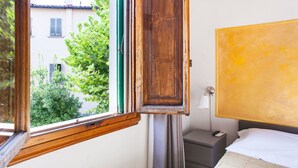 Una habitación con ventana de madera, una cama con ropa de cama blanca, una mesita de noche, una lámpara y una pared amarilla.
