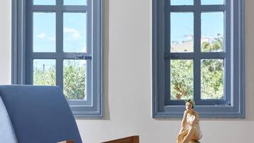 A room with a blue armchair and a small wooden table with a decorative item. Two windows with blue frames are visible.