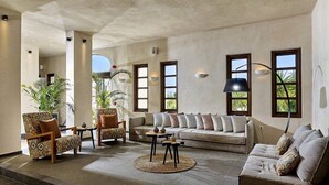 Un salón amplio con un sofá, sillones y una mesa de centro. Grandes ventanas ofrecen luz natural y vistas al verdor exterior.