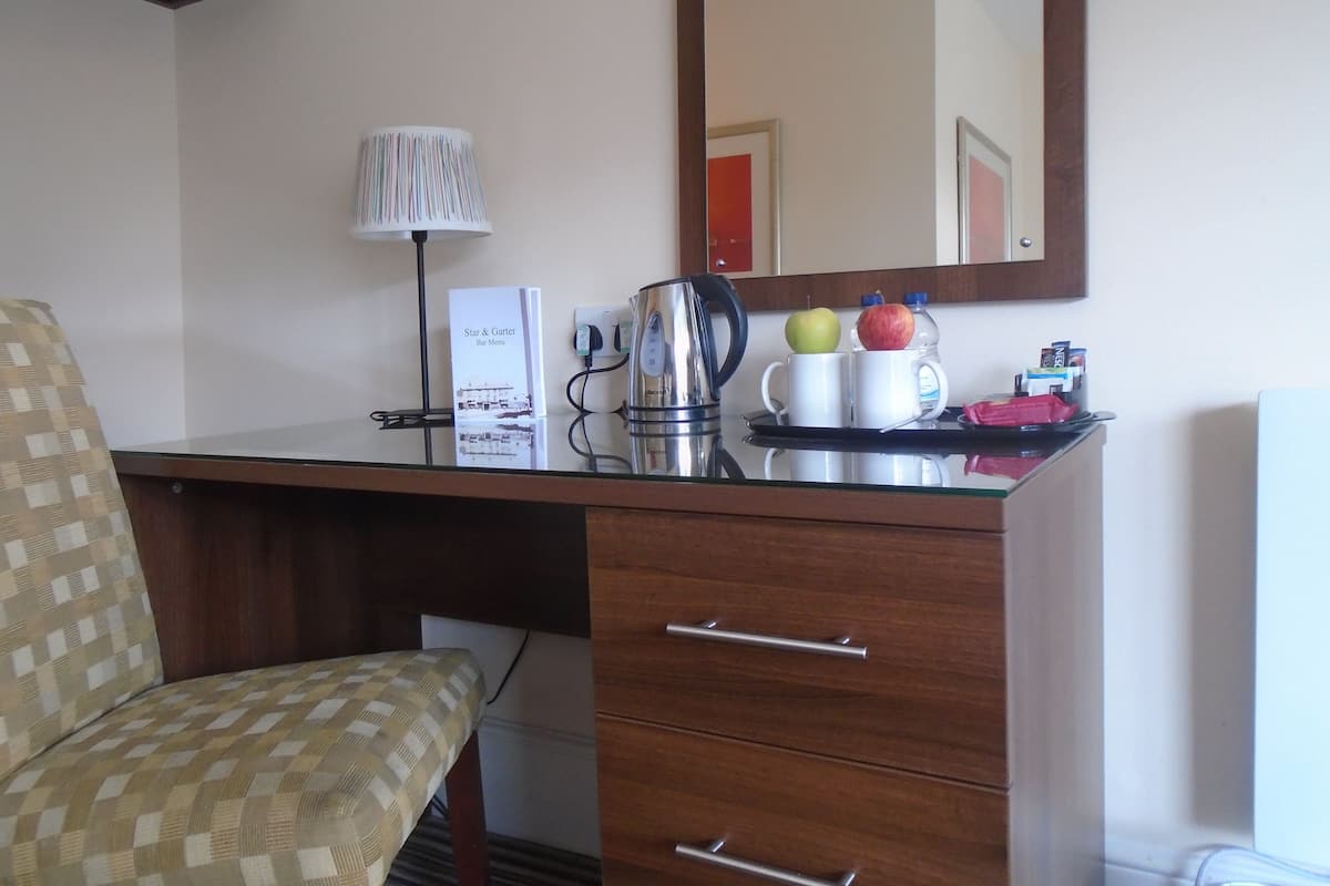 A hotel room with a wooden desk, a lamp, a kettle, two mugs, and a fruit bowl with apples.