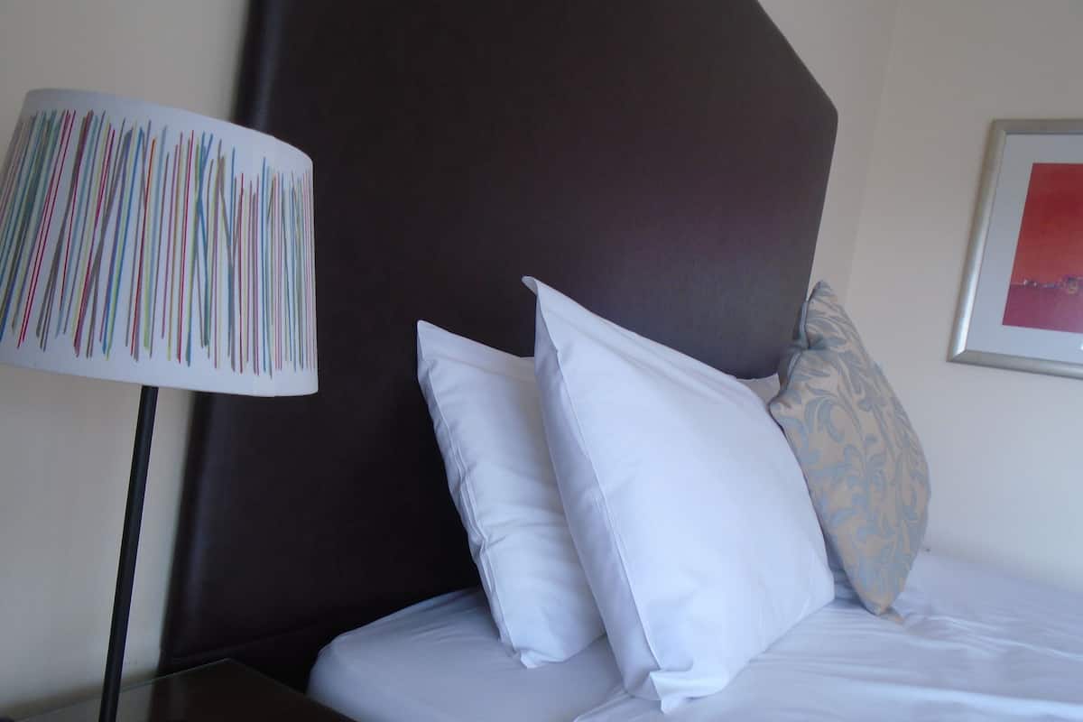 A neatly made bed with white linens, a dark headboard, and a single bedside lamp with a colorful striped lampshade.