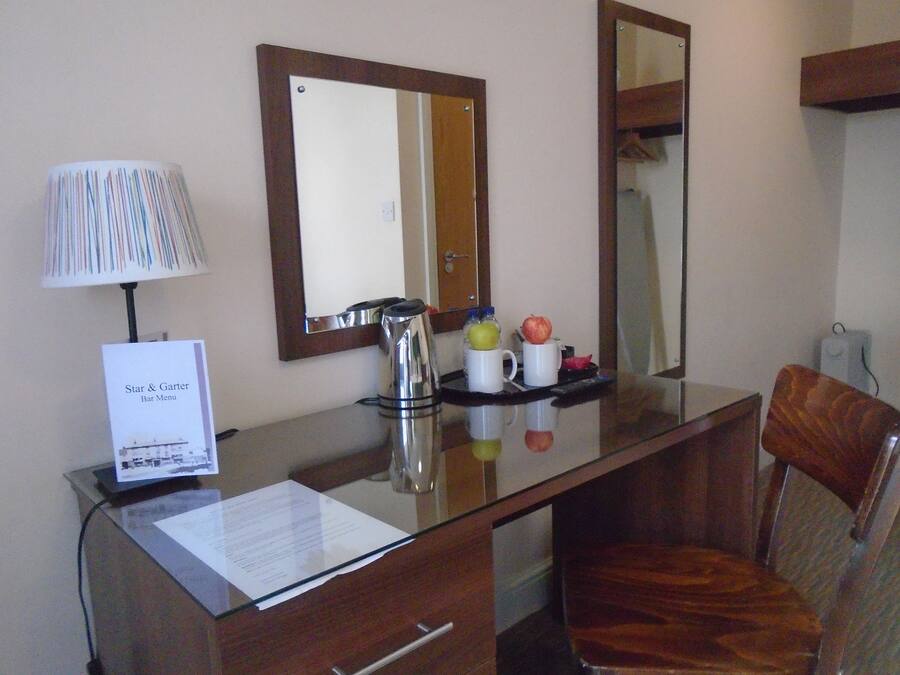 Quarto de hotel com escrivaninha, candeeiro, espelho e cadeira. Há um sinalizador na escrivaninha e uma bandeja com maçãs e uma garrafa.