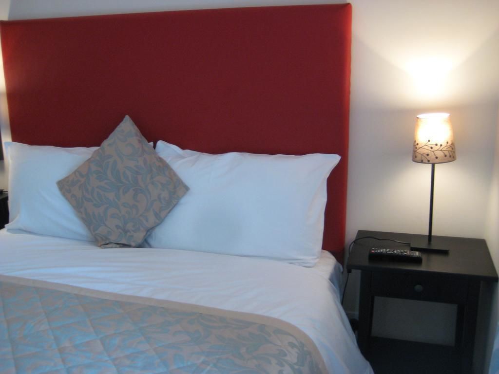 Un lit bien fait, avec une literie blanche et une tête de lit rouge, une lampe de chevet et une petite table sur laquelle se trouve une télécommande.