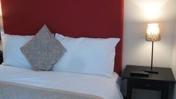 Una cama bien hecha con ropa blanca y un cabecero rojo, una lámpara de noche y una mesita con un control remoto.