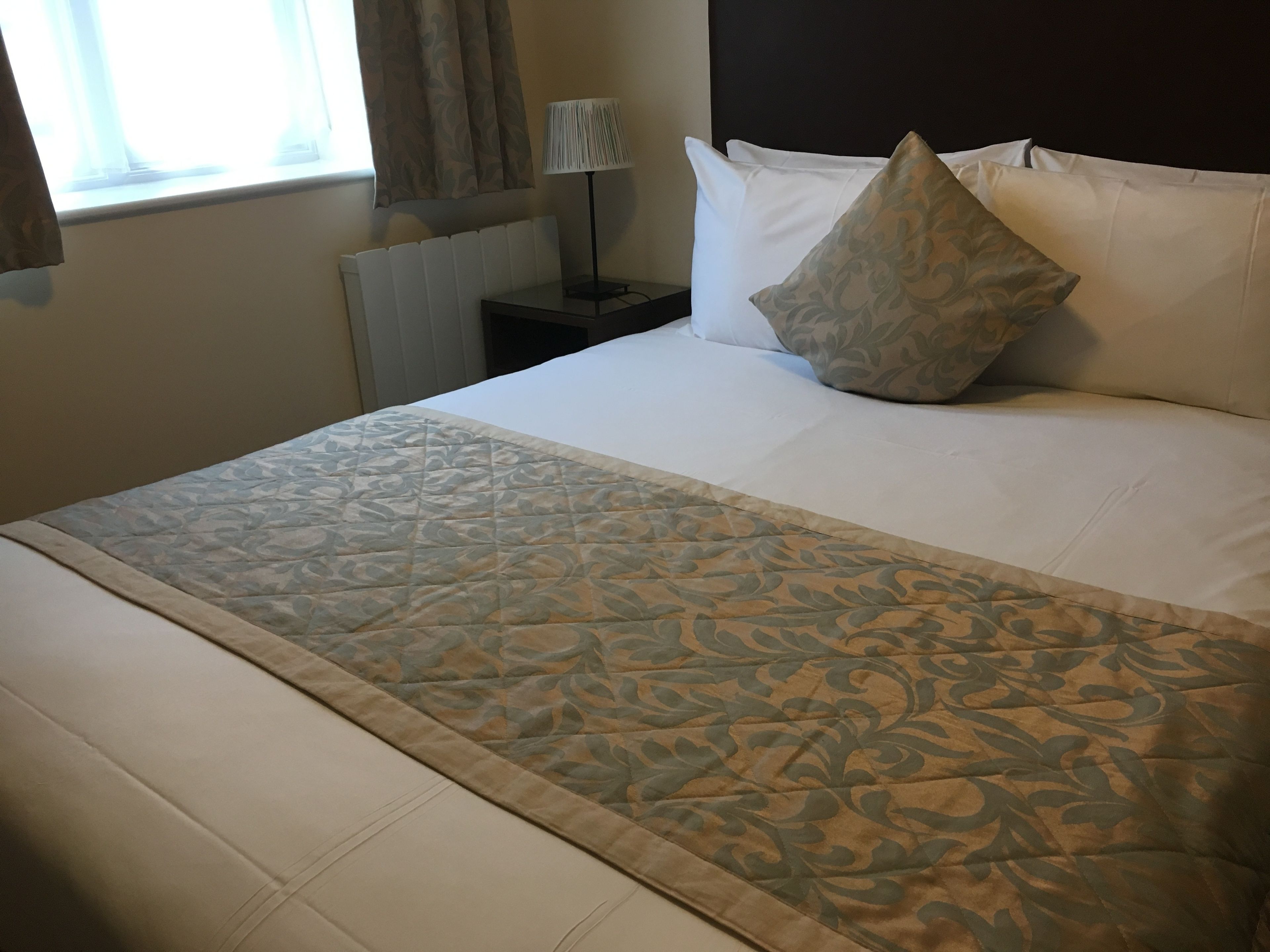 Een net opgemaakt bed met een bedsprei met patroon, een bedlampje en een hoofdbord.