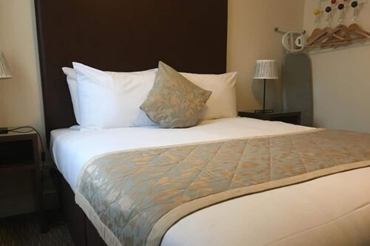A hotel room with a bed, pillows, a bedside table, a lamp, and a wall-mounted coat rack.