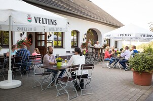 Un café al aire libre con mesas y sillas, un edificio blanco con la señalización 'VELTINS' y plantas en macetas.
