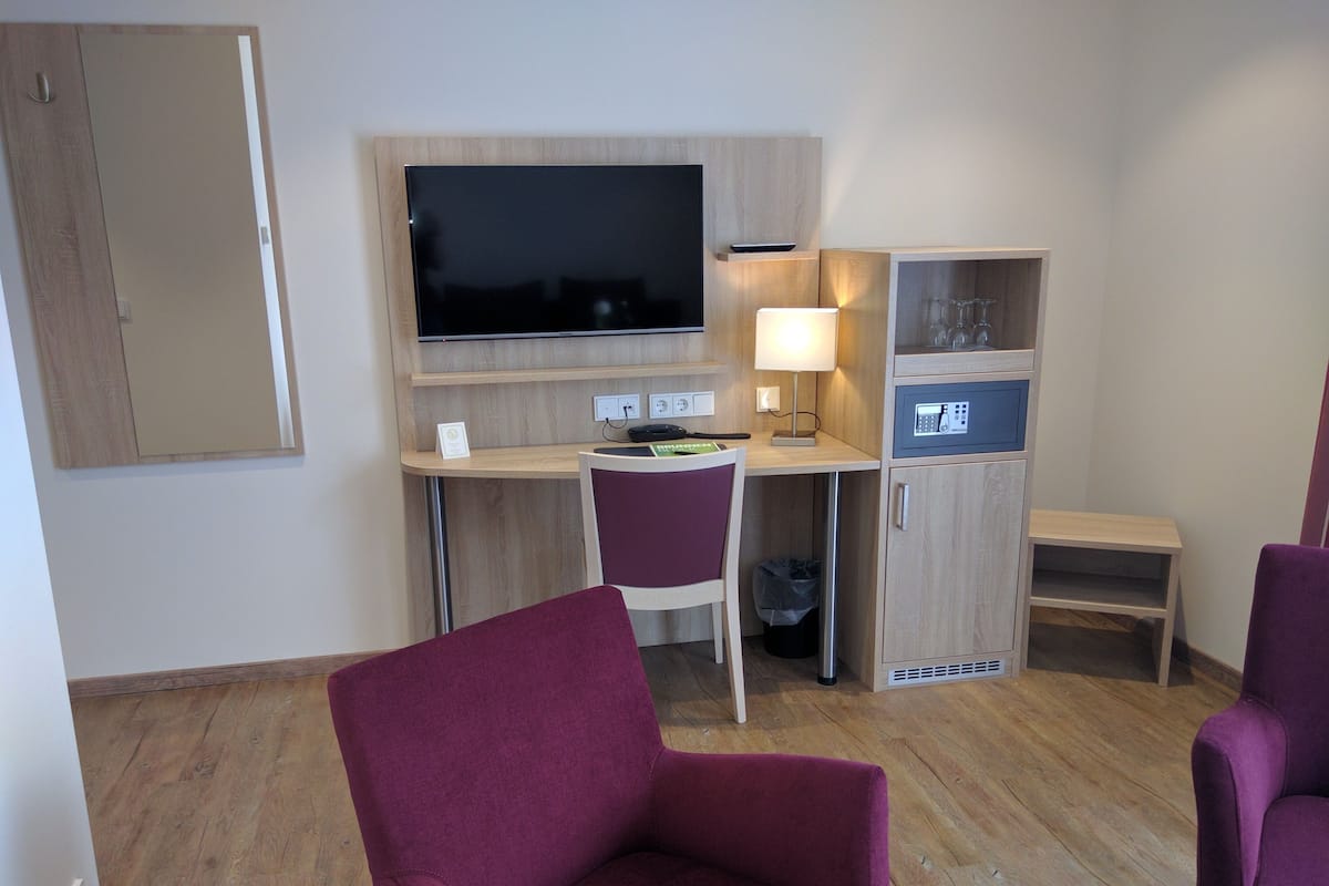 A hotel room with a desk, a television, a chair, a cabinet with a lock, and a small table.