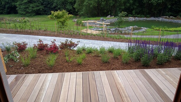 Une terrasse en bois offrant une vue sur un jardin aménagé, avec différentes espèces de plantes et un petit étang.
