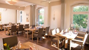 Une salle à manger bien éclairée, dotée de nombreuses tables et chaises, de grandes fenêtres offrant une vue sur la verdure et d’un plafond au design moderne.
