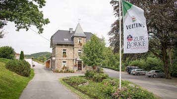 Um edifício de pedra com telhado inclinado, uma bandeira com 'VELTINS' e 'ZUR BORKE GASTSTÄTTE', carros estacionados e uma estrada que leva ao edifício.
