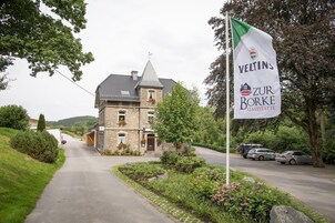 A stone building with a steep roof, a flag with 'VELTINS' and 'ZUR BORKE GASTSTÄTTE' on it, parked cars, and a road leading to the building.