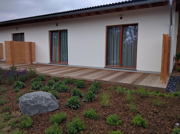 A modern house with a wooden deck, large sliding glass doors, and a garden with various plants and a rock. - Hotel Zur Borke (Neuenrade)