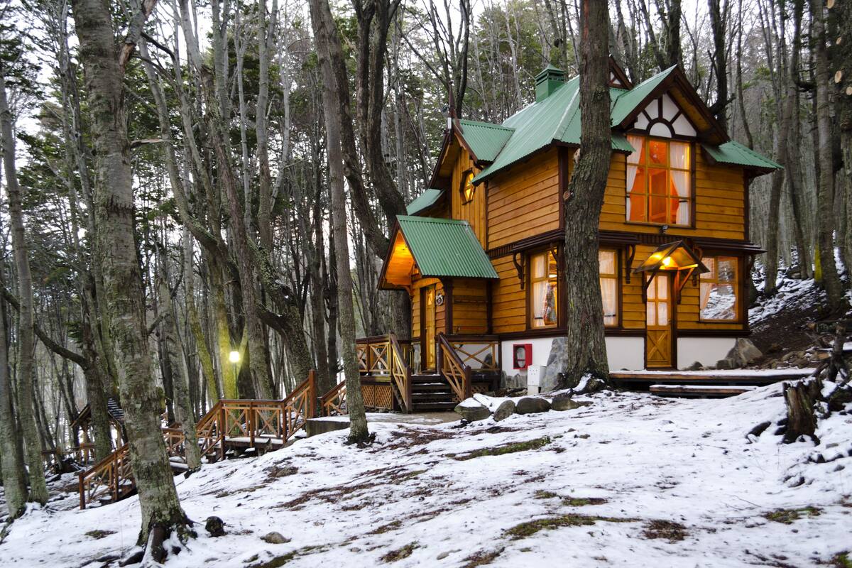 A wooden cabin with a green roof surrounded by tall trees and covered in snow.