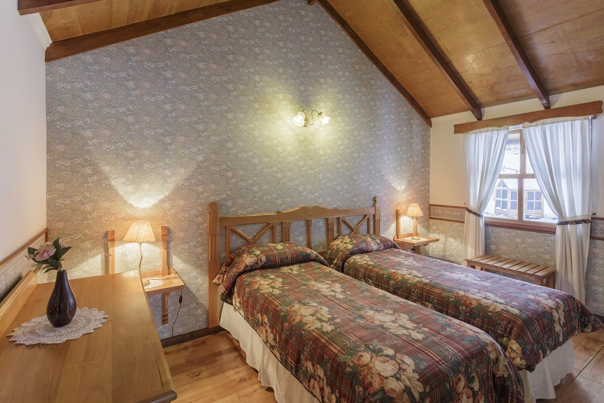 A bedroom with two beds, wooden headboards, a nightstand with a lamp, a wooden table with a vase, and a window with curtains.