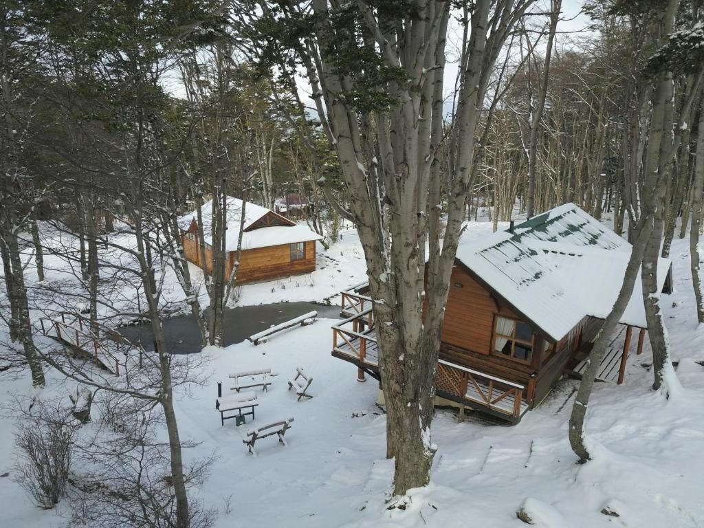 A cabin with a wooden deck and a snow-covered roof, surrounded by trees in a snowy landscape.