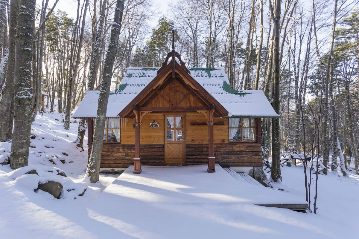 A wooden cabin with a snow-covered roof, surrounded by a forest.