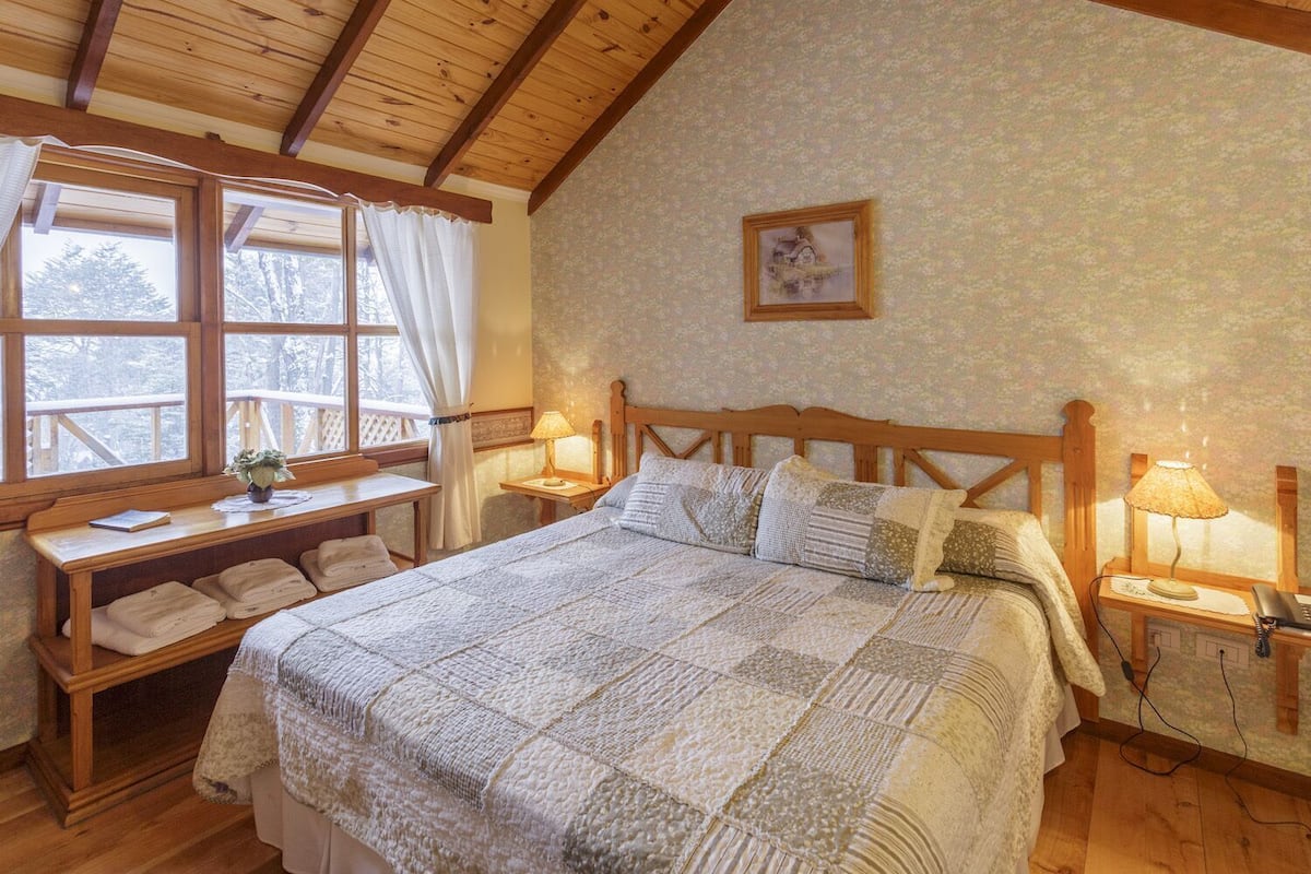 A cozy bedroom with a wooden bed, bedside lamps, a desk with a chair, and a window with curtains.