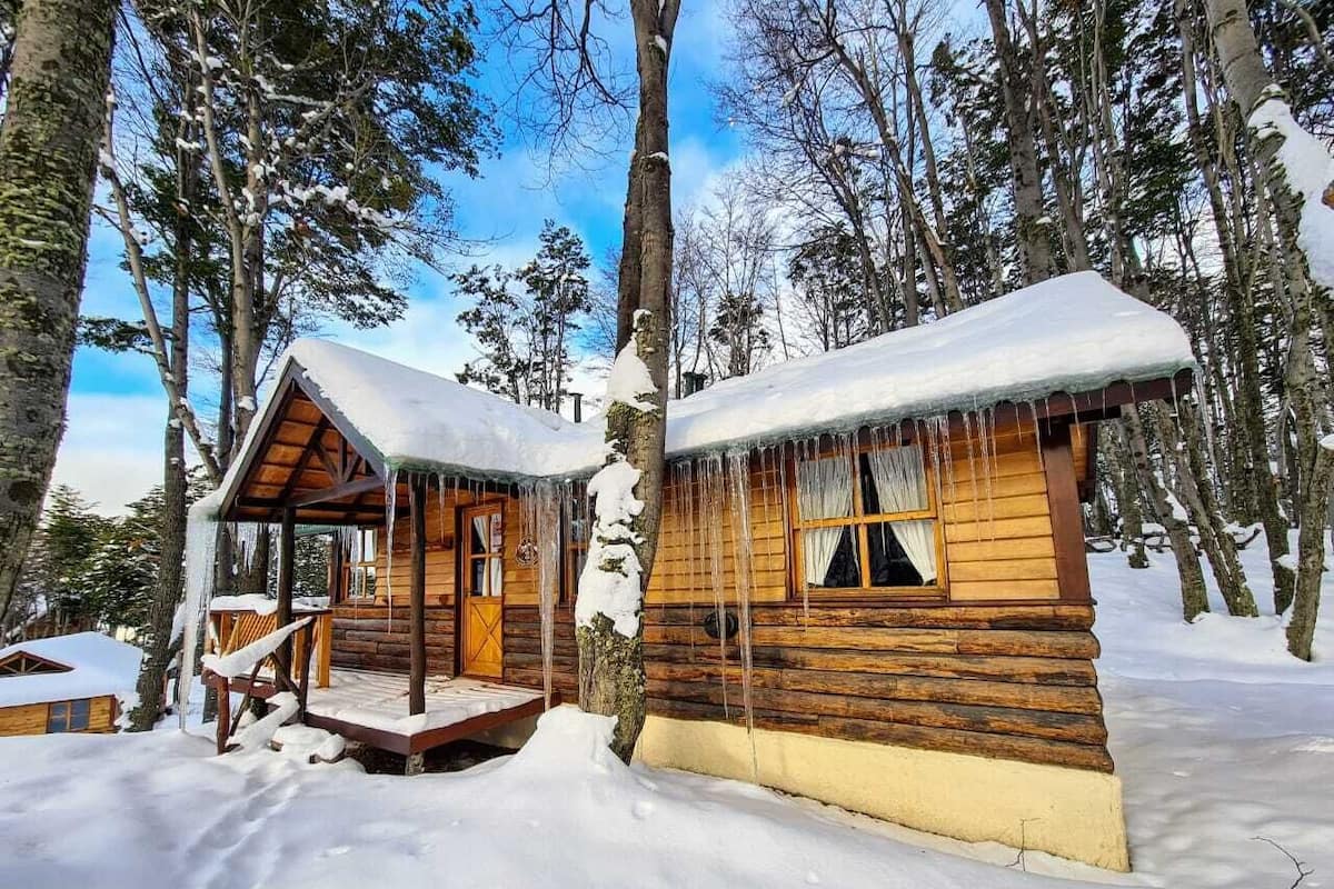 Two wooden cabins with snow-covered roofs and icicles hanging from the eaves, surrounded by trees and snow.