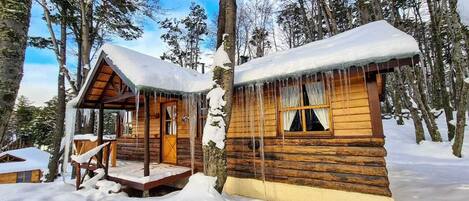 Deux cabanes en bois aux toits enneigés, avec des glaçons pendants des avant-toits, entourées d’arbres et de neige.