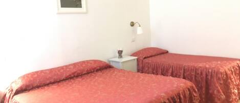 Quarto com duas camas cobertas por edredons estampados em vermelho, uma pequena mesa de cabeceira com abajur e um quadro na parede.