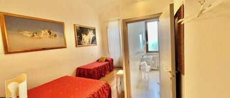 A hotel room with two beds, a desk with a lamp, a painting, and a bathroom visible through an open door.
