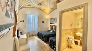 A room with two beds, a desk, a chair, a window with curtains, a bathroom with a sink and mirror, and a decorative ceiling.