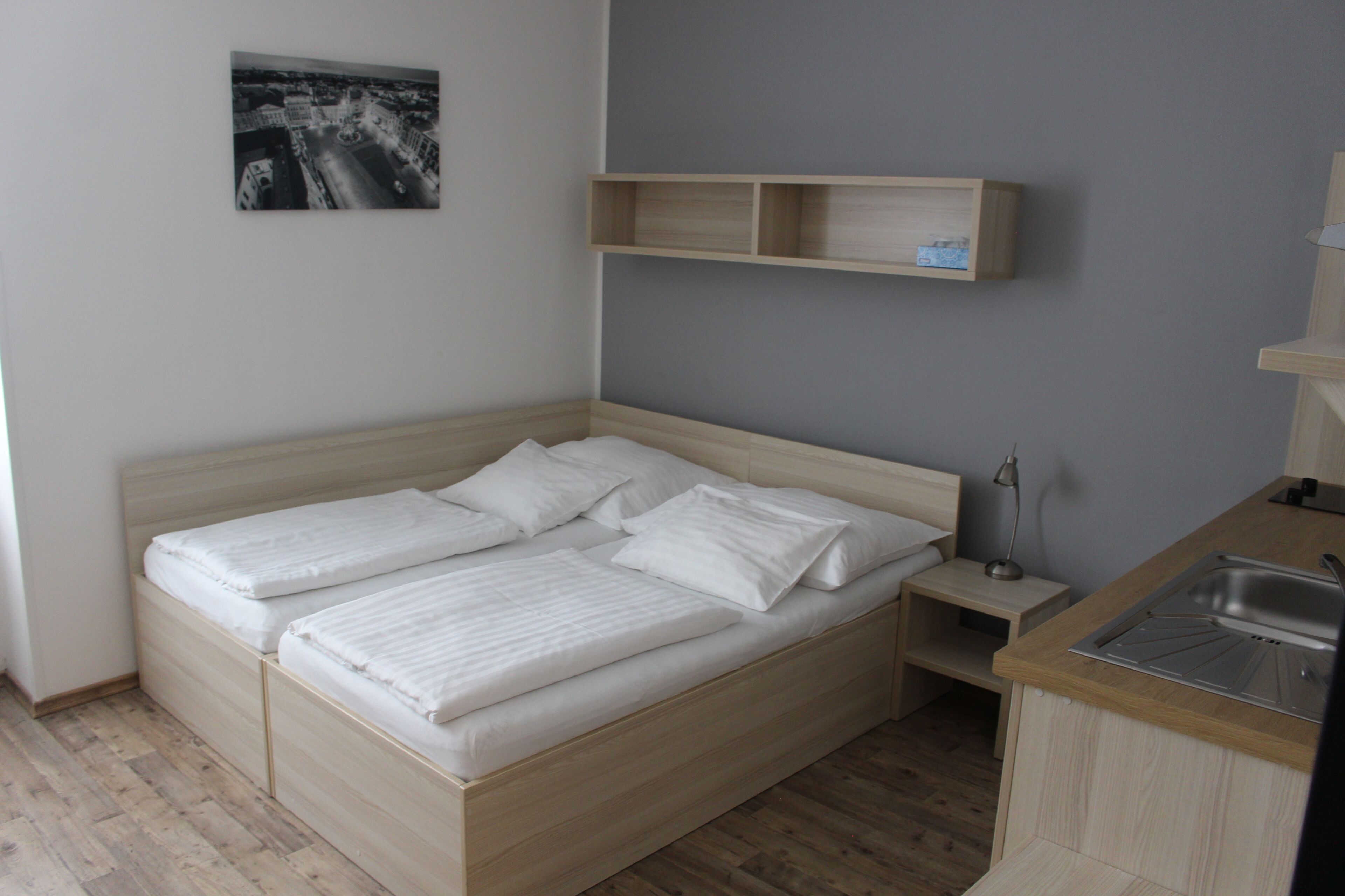 Un lit simple avec un cadre en bois et du linge de lit blanc, une petite table de chevet avec une lampe, et un coin cuisine intégré avec un évier.