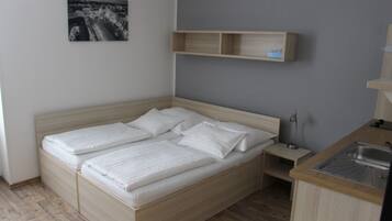 Un lit simple avec un cadre en bois et du linge de lit blanc, une petite table de chevet avec une lampe, et un coin cuisine intégré avec un évier.