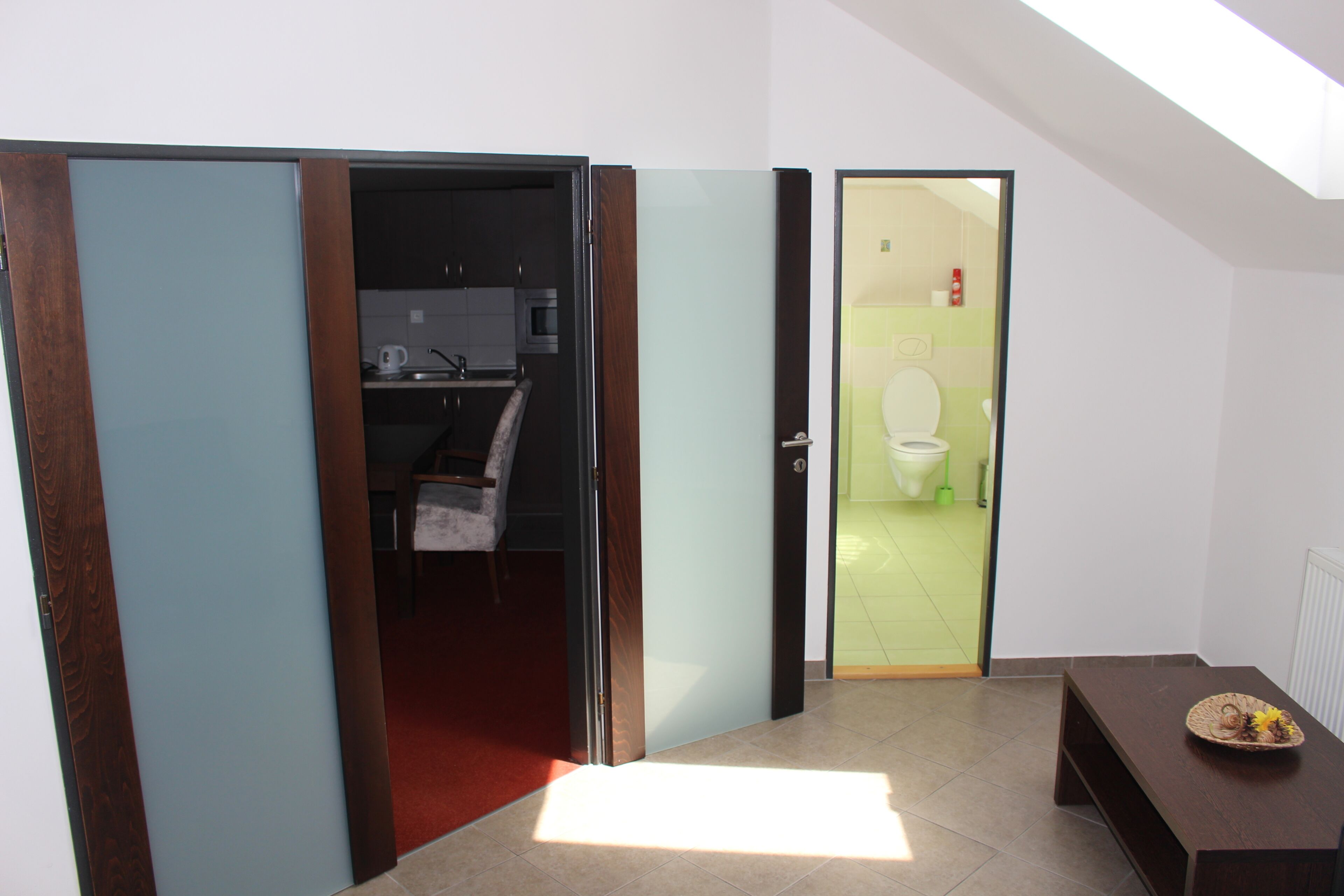 A room with a wooden floor, a bench, a door leading to a bathroom, and a kitchen area visible through an open door.
