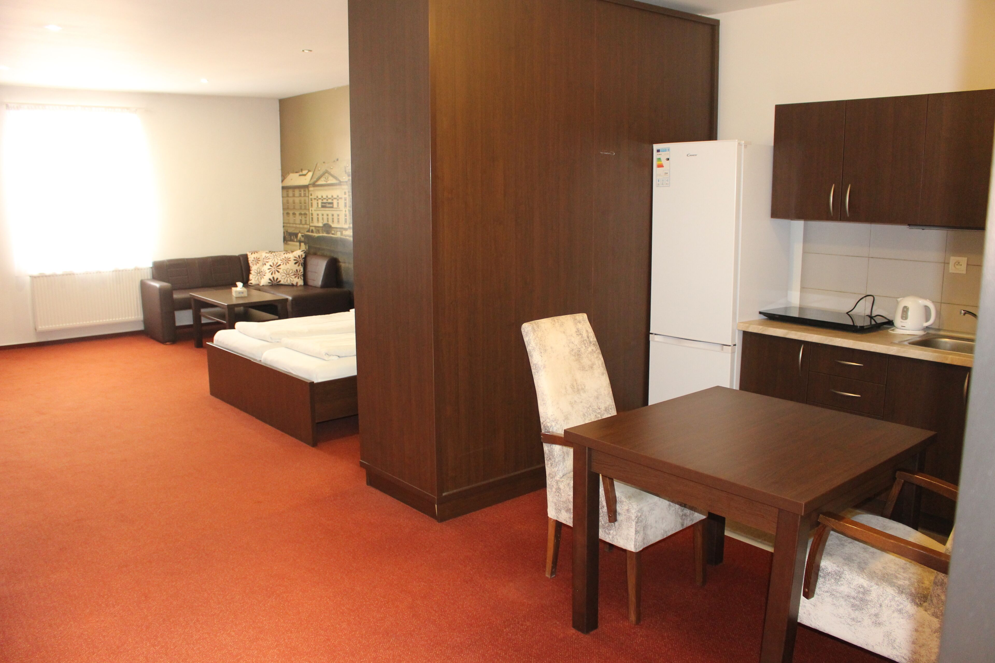 A compact hotel room with a bed, a small sofa, a dining table with chairs, a kitchenette, and a large window.