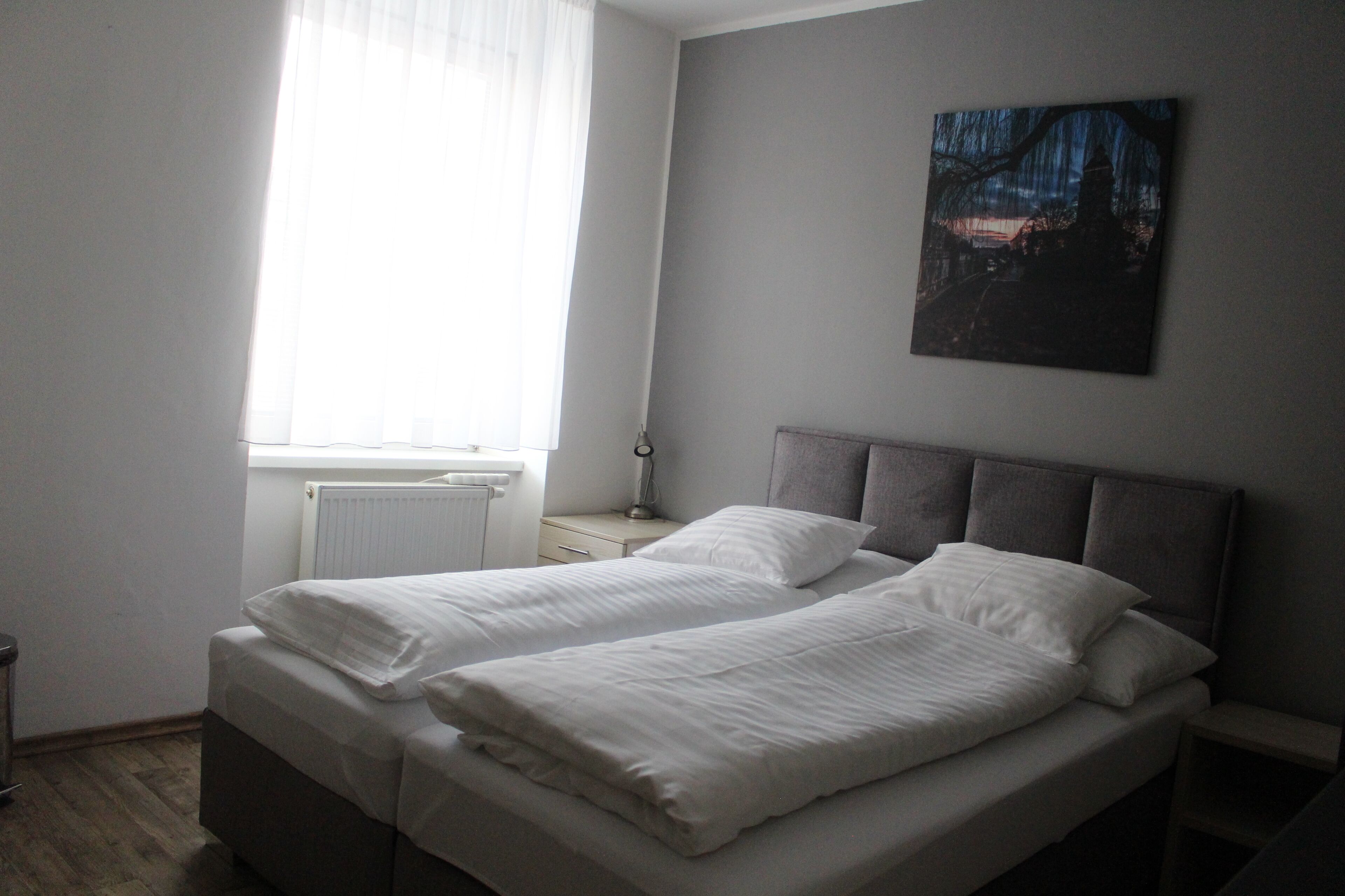 Un dormitorio con cama, mesita de noche, lámpara, ventana con cortinas y un cuadro en la pared.