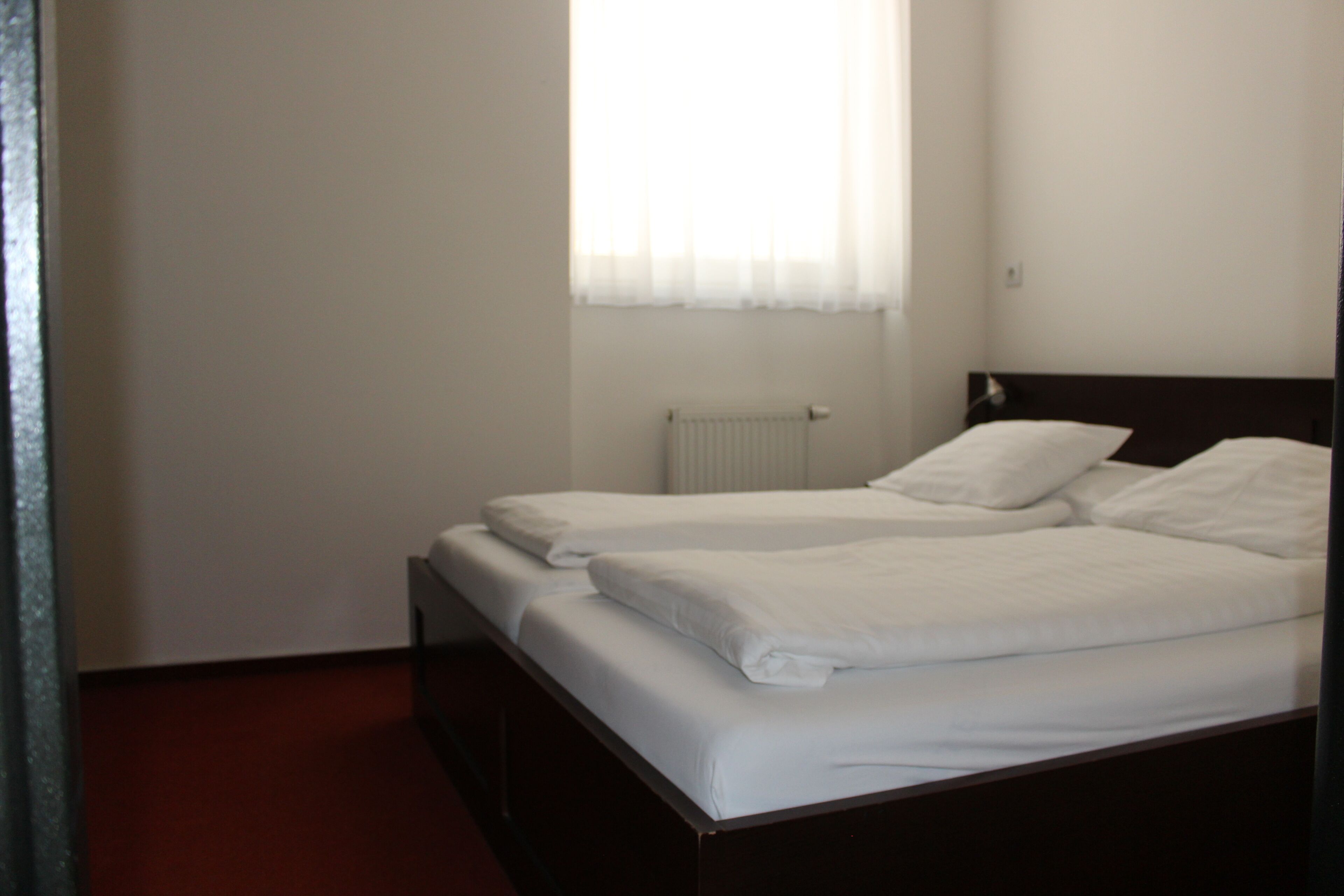 Una cama individual con ropa blanca en una habitación que tiene un radiador y una ventana con cortinas translúcidas.