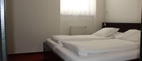 Una cama individual con ropa blanca en una habitación que tiene un radiador y una ventana con cortinas translúcidas.