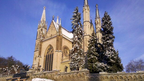 A historic church with tall spires and a large arched window, surrounded by snow-covered trees and a stone wall. - Hotel Pinger (Remagen)