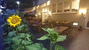 Un espacio de comedor al aire libre con mesas y sillas, un girasol y un edificio con ventanas y una puerta al fondo.