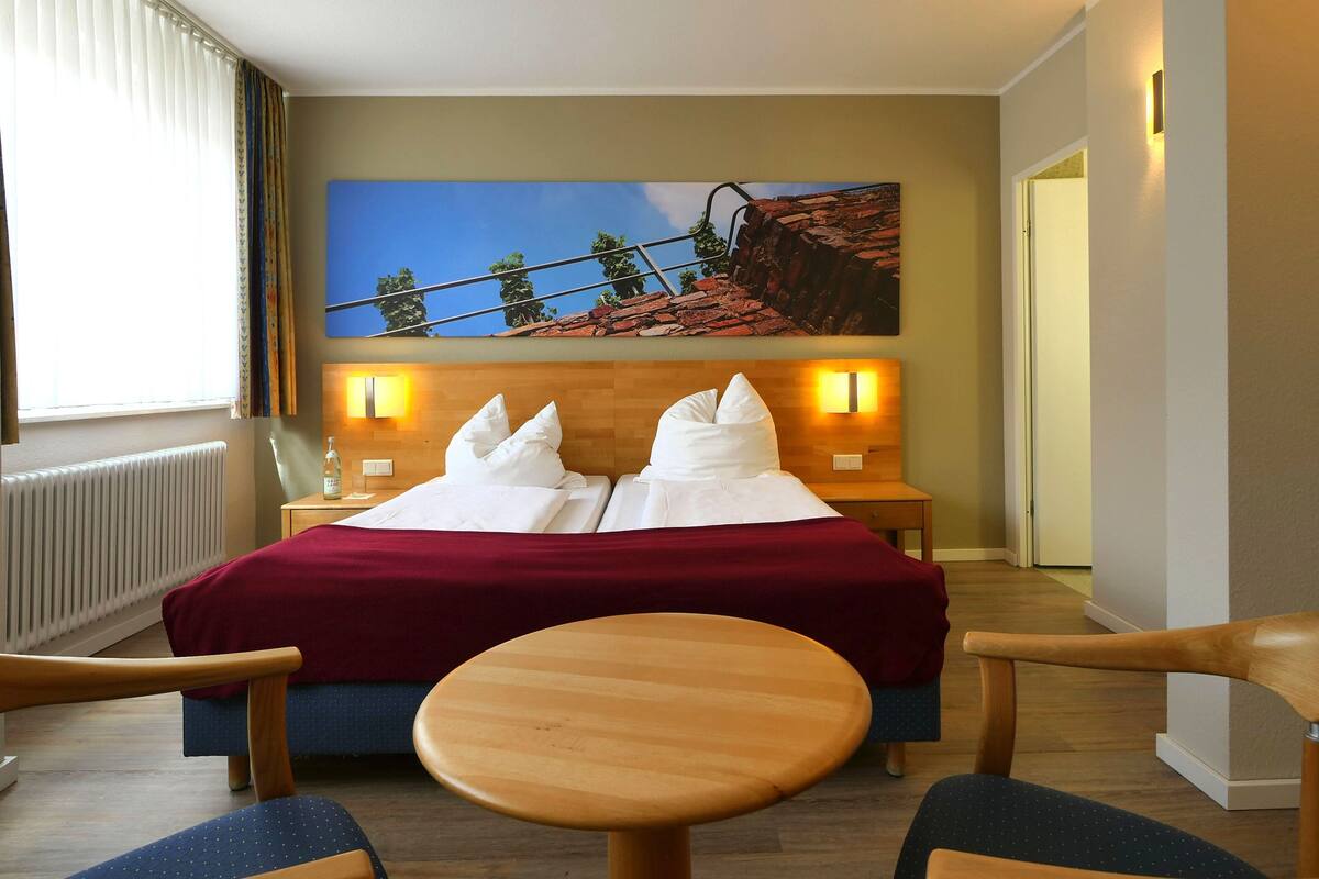 Une chambre d’hôtel avec un grand lit, deux tables de chevet avec des lampes, une tête de lit en bois, un tableau représentant un paysage et une petite table ronde avec deux chaises.