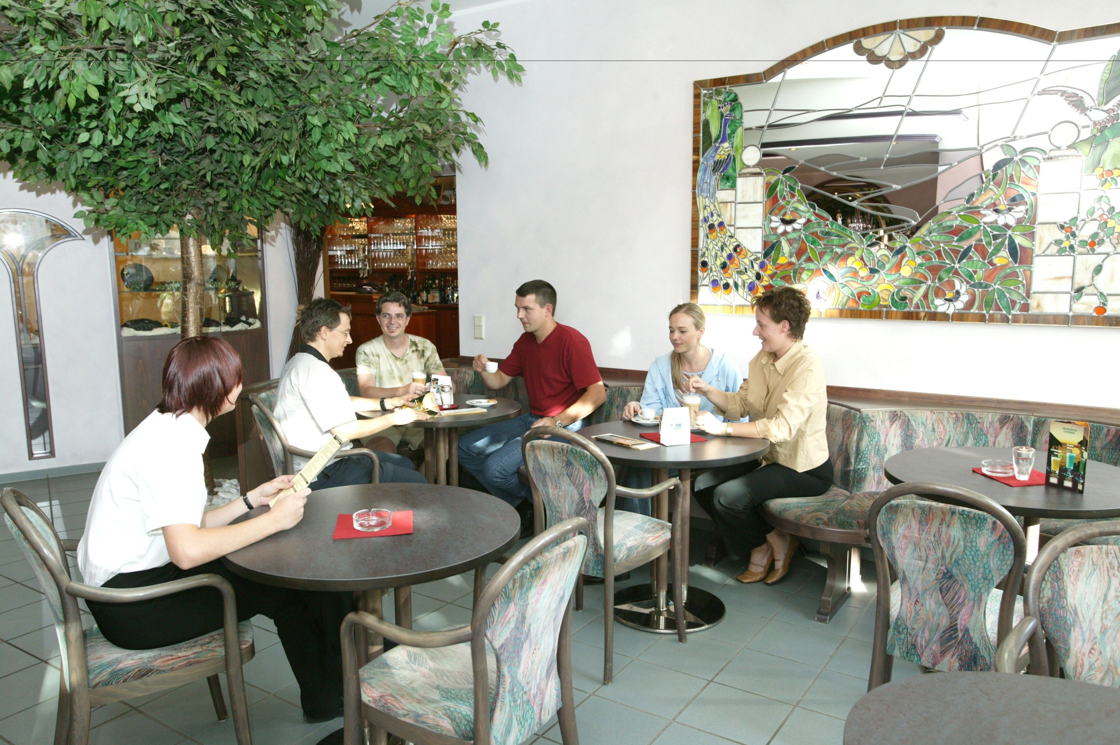 A group of people seated at round tables in a cafe with a large decorative mirror and a plant in the background.