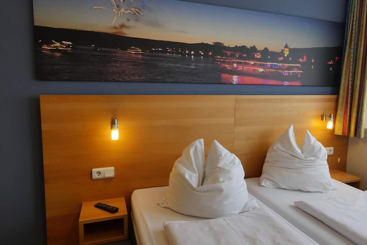 Une chambre d’hôtel avec une tête de lit en bois, deux oreillers blancs, une table de chevet avec une télécommande et un tableau représentant un coucher de soleil sur une ville.