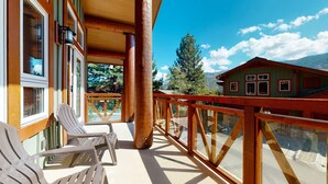 A wooden balcony with a view of a mountain and a house. There are two white chairs on the balcony.