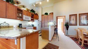 A modern kitchen with granite countertops, wooden cabinets, and a central island. There is a dining area with a table and chairs, and a hallway leading to another room.
