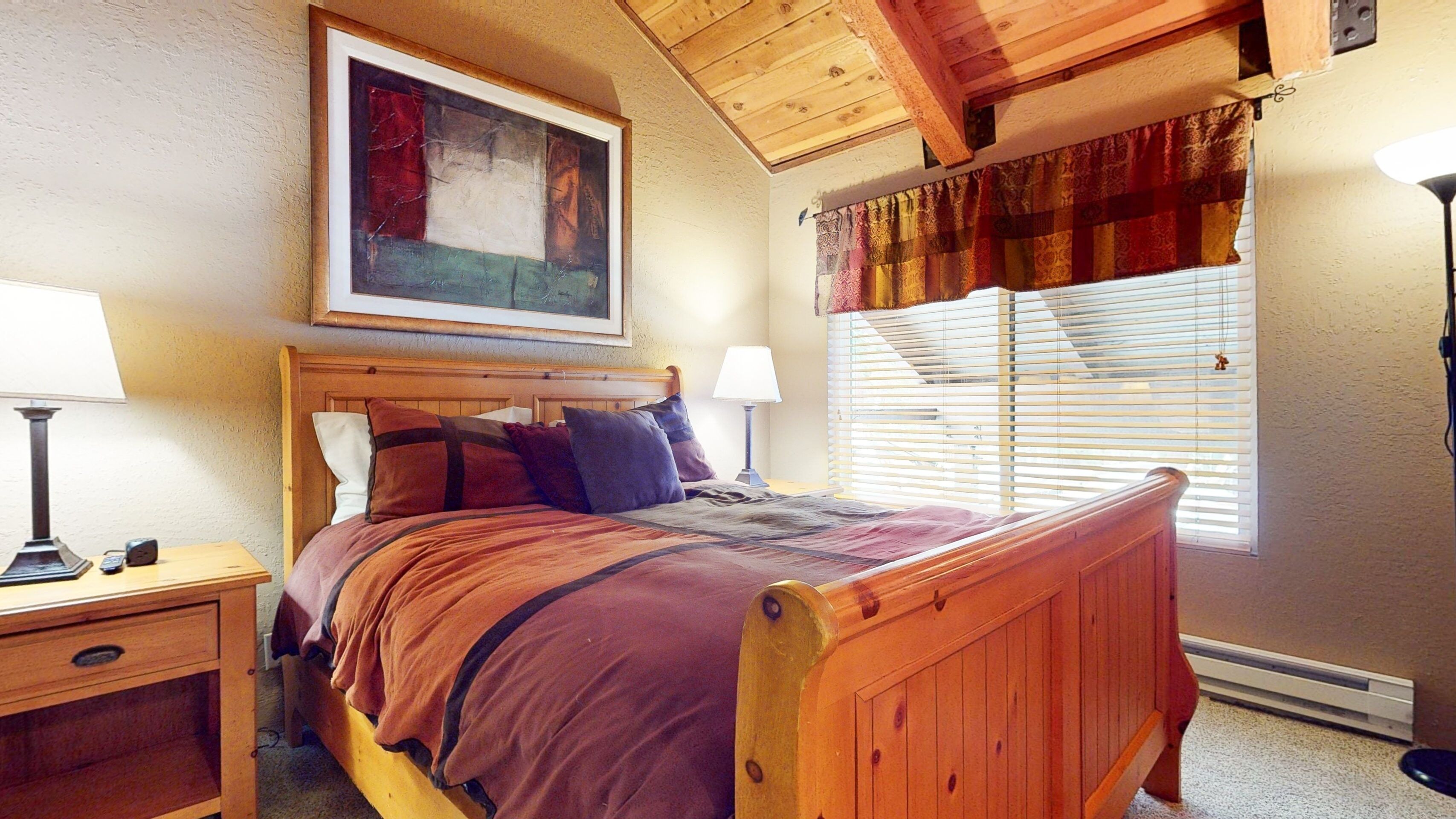A bedroom with a wooden bed, a nightstand, a lamp, a painting on the wall, and a window with curtains.