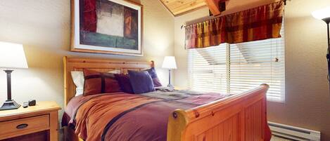 A bedroom with a wooden bed, a nightstand, a lamp, a painting on the wall, and a window with curtains.