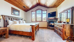 A bedroom with a wooden bed, a stone fireplace, and large windows. - Graystone 2088 4 Bedroom Home by RedAwning (Mammoth Lakes)