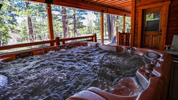 A hot tub with a wooden deck and large windows overlooking a forest. - Graystone 2088 4 Bedroom Home by RedAwning (Mammoth Lakes)