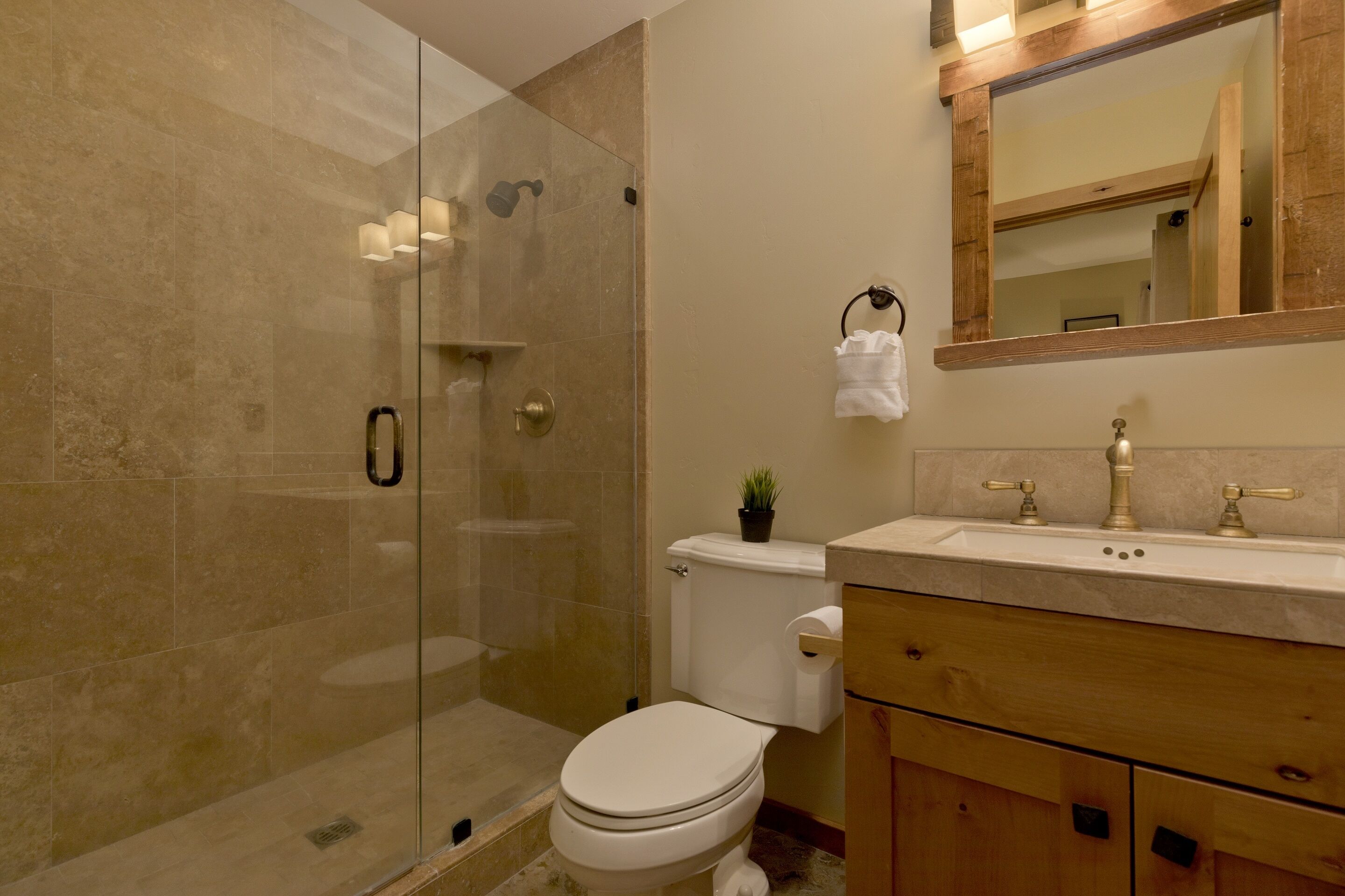 A bathroom with a glass shower enclosure, a toilet, a sink with a mirror above it, and wooden cabinets.