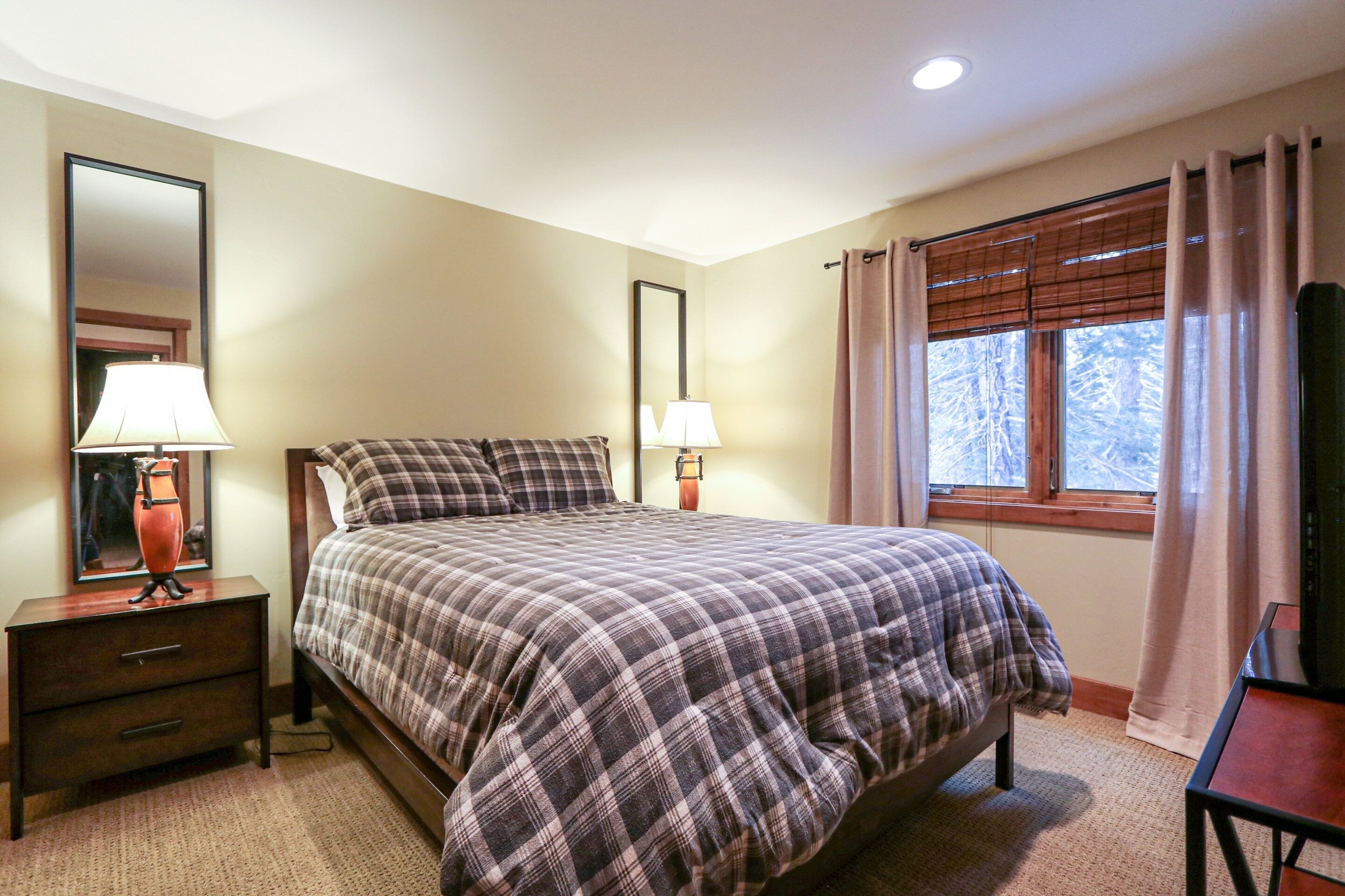A bedroom with a bed, two bedside tables with lamps, a dresser, a window with curtains, and a mirror on the wall.