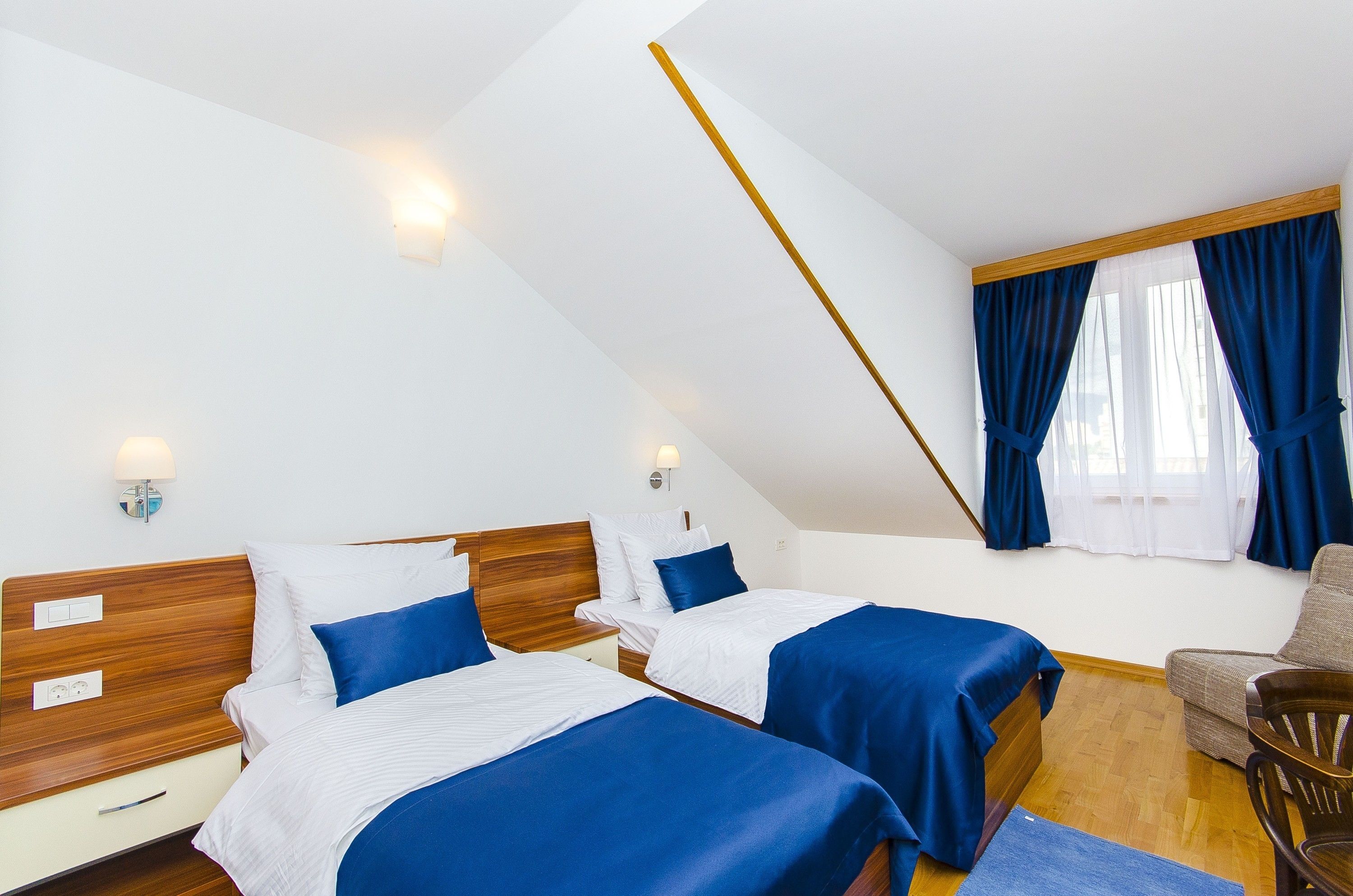 A room with two beds, wooden headboards, a wooden floor, a chair, and a window with blue curtains.